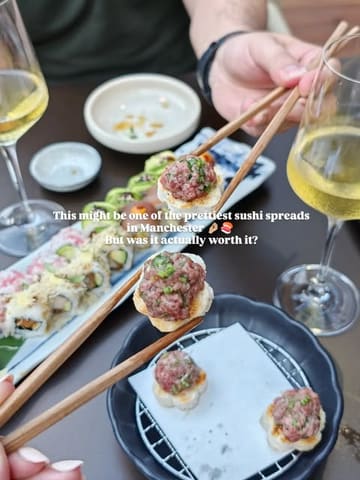 A high-quality, well-composed shot of a sushi spread at Sticks'n'Sushi, featuring a close-up of beef tataki on toast being held with chopsticks, with wine glasses and a blurred sushi platter in the background.