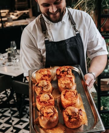 A professional, well-lit portrait of a chef presenting a tray of freshly baked, golden-brown pies, capturing the essence of a comfort food festival. The image effectively uses depth of field to keep the focus on the product while providing context of the venue.