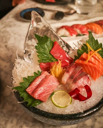 A high-quality, close-up shot of a premium sashimi platter served on a bed of ice, featuring various cuts of raw fish, garnished with shiso leaves and citrus. The lighting is warm and inviting, highlighting the fresh texture of the fish.
