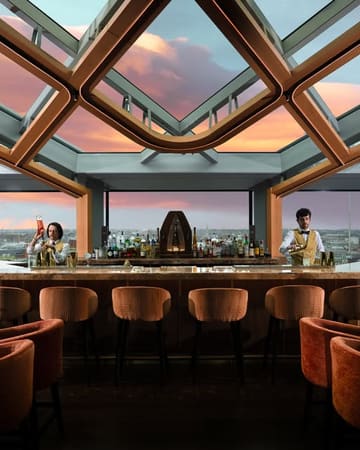 A highly polished, symmetrical shot of a bar interior featuring a glass ceiling and a sunset view over the city. The image creates an aspirational atmosphere, positioning the venue as an upscale destination for evening drinks.