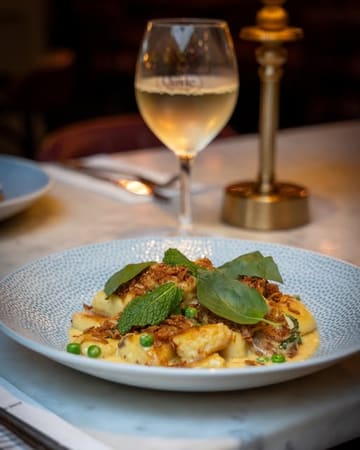 A high-quality, professional food photograph featuring a plate of homemade Parmesan gnocchi served with garden peas, shallot velouté, fresh basil, mint, and crispy shallots, accompanied by a glass of white wine in a softly blurred background.