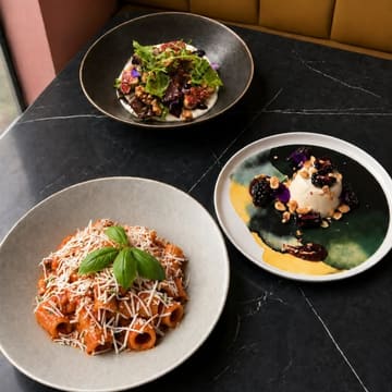A high-quality, overhead food photography shot featuring three distinct dishes—a salad, a pasta dish, and a dessert—arranged on a dark marble table, promoting an early evening menu at a restaurant.