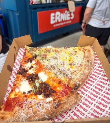 A high-quality, close-up shot of two slices of pizza served in a branded takeaway box. The lighting is natural and bright, highlighting the textures of the cheese, chili, and crust, making the food look fresh and appealing. The composition is tight and focused on the product, effectively showcasing the quality of the pizza.