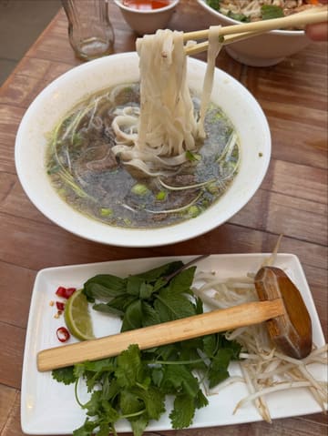 A top-down/45-degree shot of a bowl of beef Pho being lifted with chopsticks, accompanied by a side plate of fresh herbs, bean sprouts, lime, and chili on a wooden table.