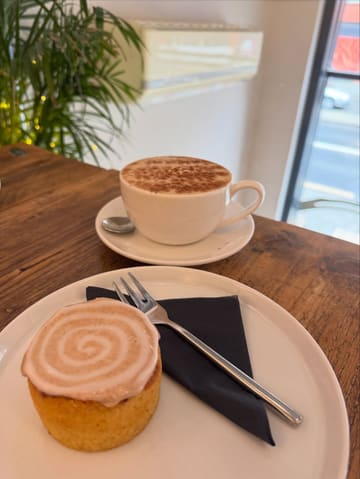 A simple, natural-light shot of a coffee and a cinnamon roll served on a wooden table, likely in a cafe.