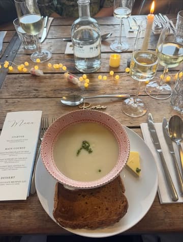 A top-down, atmospheric shot of a wedding meal setting, featuring a bowl of soup and a slice of bread on a rustic wooden table. The scene is decorated with fairy lights and includes a printed menu, wine glasses, and place settings.