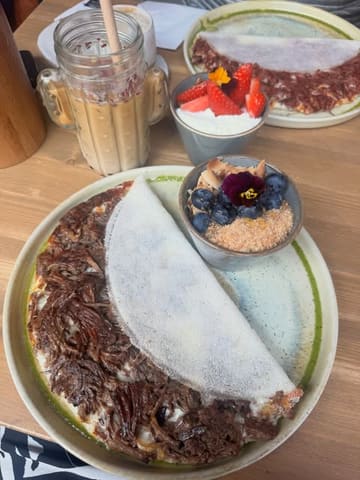 A top-down, natural light shot of a brunch spread featuring a beef brisket tapioca crepe, an acai bowl, and an iced latte. The image highlights a casual dining experience with a focus on gluten-free options.