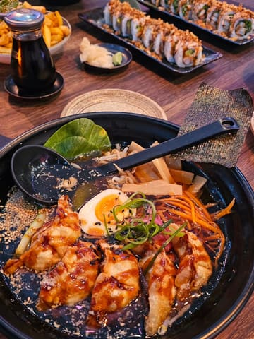 A top-down, casual shot of a ramen bowl featuring gyoza, with a side of fries and a plate of sushi rolls visible in the background on a wooden table.