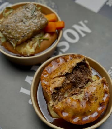 A top-down shot of two hearty pub-style dishes served in disposable bowls on a branded table. One bowl features a golden-brown pie with a flaky crust, while the other contains a Yorkshire pudding filled with mash, gravy, and meat. The lighting is soft and natural, highlighting the textures of the food.