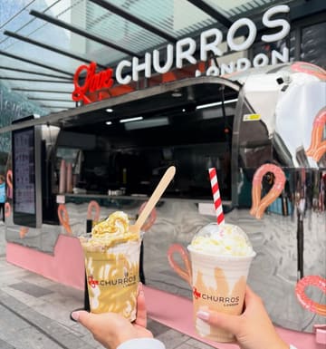 A bright, clear, and well-composed shot of a pistachio sundae and a hazelnut milkshake held in front of a Love Churros London food truck. The image effectively highlights the products in their natural, casual setting.