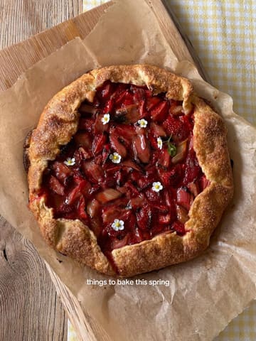 A high-quality, overhead shot of a freshly baked fruit galette, likely strawberry and rhubarb, garnished with small white flowers. The pastry is golden brown and the fruit filling looks glossy and vibrant, presented on parchment paper atop a rustic wooden board.