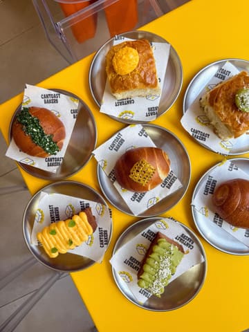 A vibrant, high-quality overhead flat-lay photograph showcasing a variety of baked goods from Cantoast Bakery, arranged on individual metal plates with branded parchment paper on a bright yellow table.