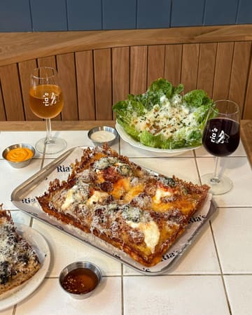 A high-quality, well-composed flat-lay photograph of a deep-dish pizza meal, accompanied by a fresh salad, dipping sauces, and glasses of wine, set against a clean, warm-toned wooden background.