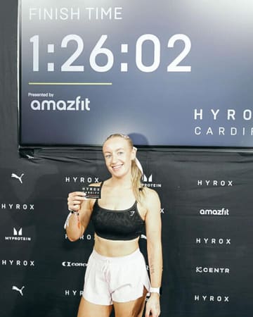 A photo of a participant at a Hyrox fitness event, standing in front of a branded backdrop displaying her finish time.