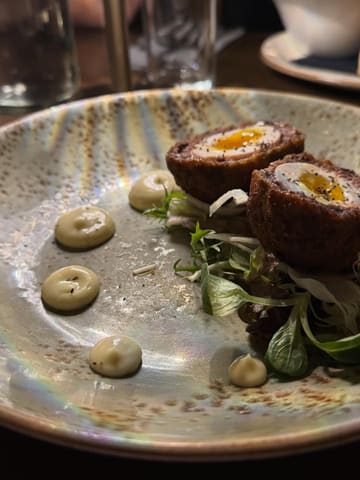 A close-up, eye-level shot of a Scotch egg starter served at a gastro pub. The dish is presented with a small side salad and dots of mustard mayonnaise on a textured, speckled plate.