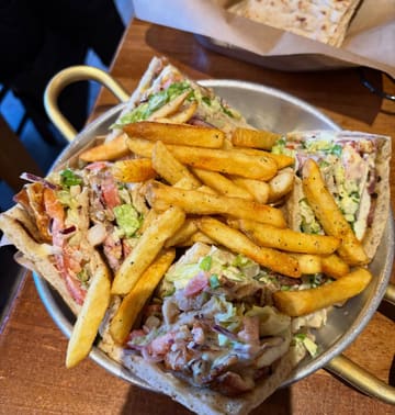 A top-down, casual shot of a Greek-style platter featuring a club sandwich, hummus, and halloumi fries, served in a metal tray on a wooden table.