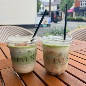 A bright, well-composed shot of two iced matcha drinks served in clear plastic cups, placed on a wooden outdoor table at a cafe.