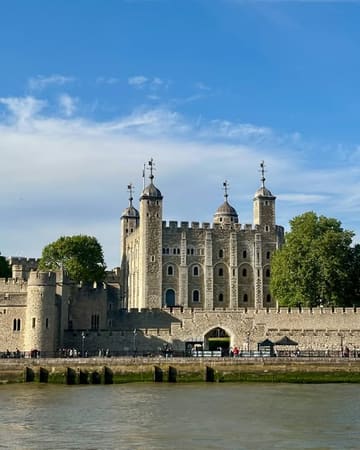A clear, well-composed photograph of the Tower of London taken from the riverbank. The image captures the historic architecture under a bright blue sky, serving as a straightforward travel or tourism shot.