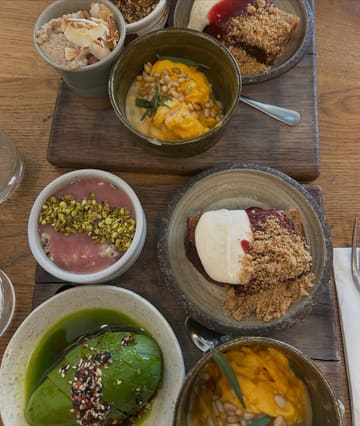 An overhead flat-lay shot of a build-your-own brunch board featuring various small bowls containing avocado, scrambled eggs, oats, and banana bread, served on wooden boards.