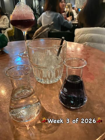 A casual, dimly lit snapshot taken at a bar, featuring a molecular-style cocktail setup with a coupe glass, a beaker, and a glass of ice. The image captures a typical night-out atmosphere.