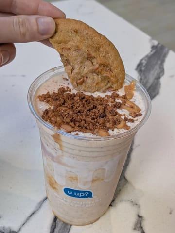 A close-up, eye-level shot of a milkshake topped with a cookie, held by a hand against a marble surface. The image is a standard, casual product shot typical of social media reviews for takeaway desserts.