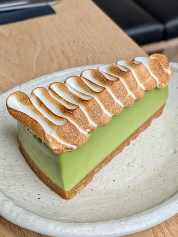 A high-quality, close-up shot of a slice of matcha cheesecake topped with a torched lime meringue, presented on a textured ceramic plate.