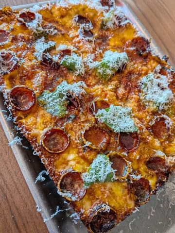 A high-quality, overhead close-up shot of a Detroit-style pizza, showcasing the crispy edges, pepperoni, and melted cheese, served on a metal tray.