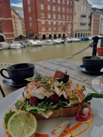 A casual, lifestyle-focused shot of a brunch dish featuring chorizo and eggs on toast, served with coffee, captured at an outdoor table overlooking a harbour.