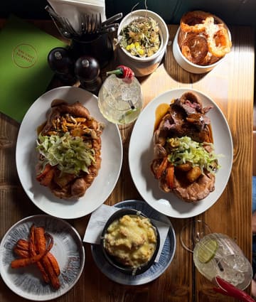 A high-quality, top-down flat lay photograph of a Sunday roast spread at a pub. The image features two large Yorkshire puddings filled with roast meats and vegetables, accompanied by sides like mac and cheese, onion rings, and roasted carrots, along with cocktails. The lighting is natural and warm, highlighting the textures of the food.