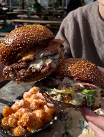 A close-up, handheld shot of two loaded smash burgers being held up in front of a pub beer garden setting, with a side of loaded fries visible in the foreground.