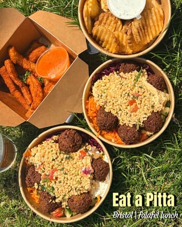 A casual, overhead flat-lay shot of a picnic spread featuring falafel boxes, halloumi fries, and waffle fries on a grass background. The image captures a relaxed, outdoor lunch setting.