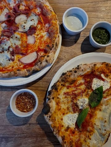 An overhead shot of two pizzas and several small dipping bowls on a wooden table, likely taken at a casual Italian restaurant. The image focuses on the food, showcasing the textures of the pizza toppings and the accompanying dips.