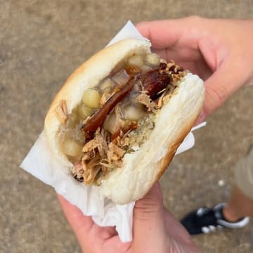 A close-up, handheld shot of a hog roast sandwich at a food market, showcasing the tender pork, stuffing, and crackling in a soft roll.