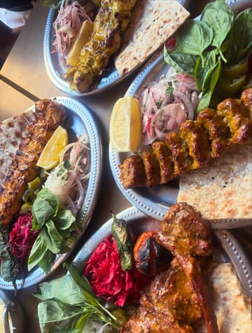 A vibrant, high-angle flat-lay photograph capturing a Persian feast, featuring multiple metal plates filled with grilled meats, flatbreads, fresh herbs, and colourful side dishes.