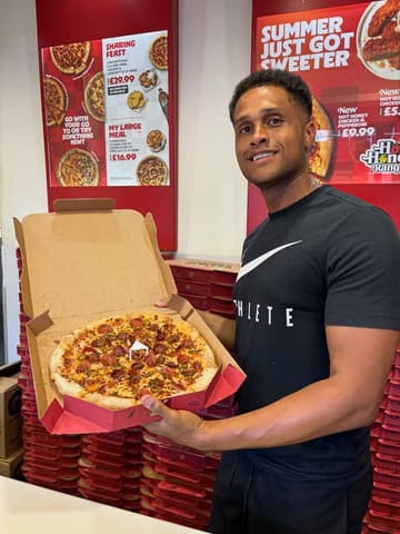 A promotional shot featuring a creator holding a pizza box in front of a Pizza Hut menu board, likely part of a behind-the-scenes or review-style video.