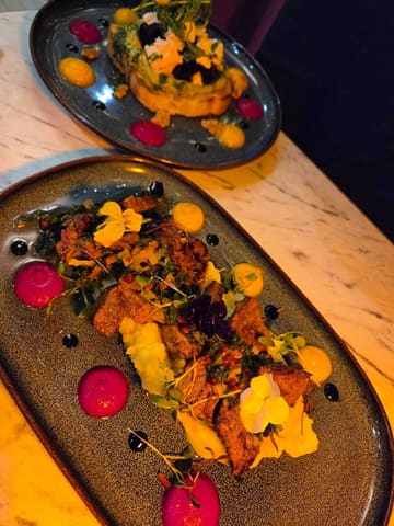 A high-angle, close-up shot of two dishes served at a restaurant. The foreground features a plate with steak, mashed potatoes, chimichurri, and edible flowers, while the background shows a plate of bruschetta with goat cheese, beetroot, and avocado.