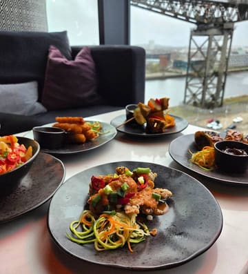 A table spread of various small plates including halloumi fries, Thai dirty fries, and chicken skewers, set against a window view of a bridge in Glasgow.