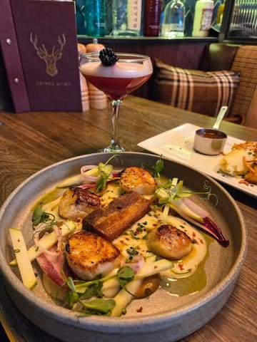 A high-quality, well-composed shot of a scallop dish served at a Scottish restaurant, accompanied by a cocktail and a menu in the background, effectively capturing the dining atmosphere.