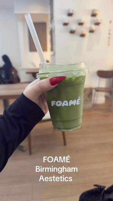 A casual, user-generated style shot of a matcha latte in a branded takeaway cup held by a customer at FOAMÉ in Birmingham. The image is functional but lacks professional polish, with flat lighting and a cluttered background.