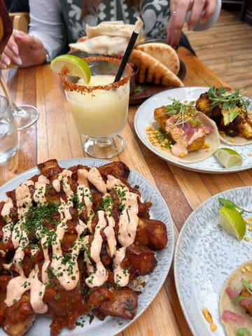 A top-down shot of a table spread featuring patatas bravas, tacos, and a cocktail. The lighting is natural but includes a distracting shadow from the photographer. The composition is slightly cramped, with plates close to the frame edges.