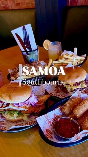 A top-down, casual table spread shot featuring various burgers, sides, and cocktails at a restaurant called Samoa. The image is clearly intended to showcase a variety of menu items in a single, indulgent feast-style shot.