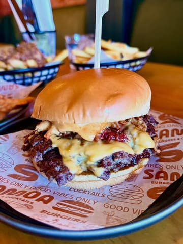 A close-up, appetizing shot of a double smash burger served on branded paper, with a basket of fries visible in the blurred background, effectively showcasing the venue's casual dining offering.