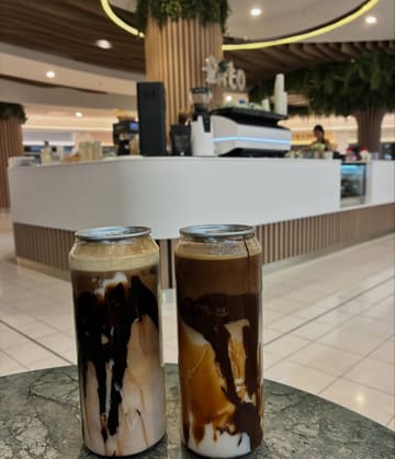 A photo of two canned iced lattes placed on a marble-effect table inside a shopping centre cafe. The drinks feature visible layers of chocolate sauce and coffee, presented in tall, clear cans.