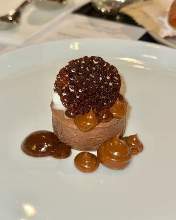 A close-up, top-down shot of a single, small-portion dessert from a tasting menu, featuring a chocolate mousse base topped with a dark tuile, served with dollops of caramel or sauce on a white plate.