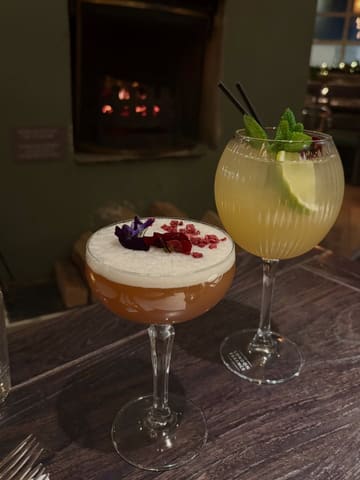 A close-up shot of two cocktails placed on a wooden table in a dimly lit pub, with a fireplace visible in the background.