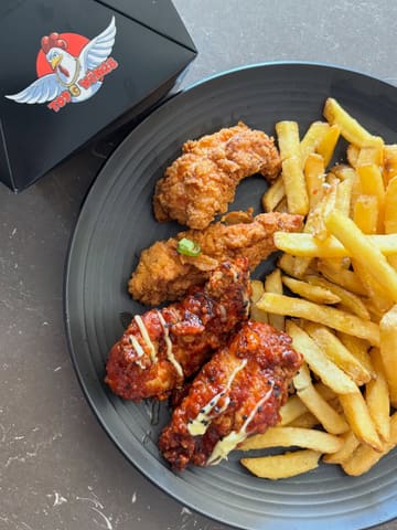 A top-down, casual shot of a plate featuring breaded chicken strips and glazed wings alongside a portion of fries, with a branded takeaway box visible in the corner.
