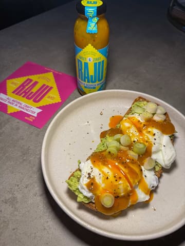 A product-focused shot featuring a bottle of 'Raju' mango and scotch bonnet sauce alongside a plate of avocado toast with poached eggs. The image serves as a promotional post for the sauce brand, highlighting its use in home cooking.
