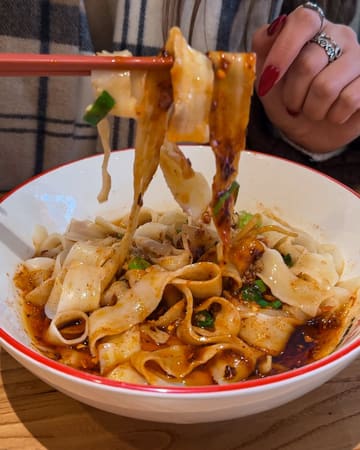A high-quality, close-up action shot of chilli oil noodles being lifted with chopsticks, effectively capturing the texture and sauce of the dish in a casual dining setting.