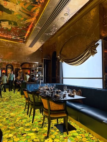 A well-composed, atmospheric interior shot of The Ivy Asia, showcasing the restaurant's distinctive, opulent decor, including the illuminated floor and ornate ceiling artwork. The image effectively captures the mood and aesthetic of the venue, making it an inviting space for diners.