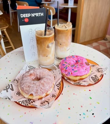 A bright, high-quality photograph featuring two iced coffees and two artisan donuts served on branded paper, showcasing a collaboration between Deep North and Mother Mercy.